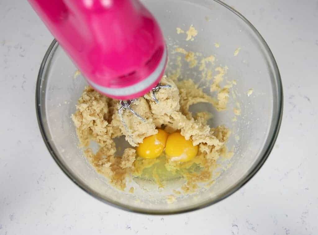 pink hand mixer creaming cookie dough, and egg and egg yolk added into bowl