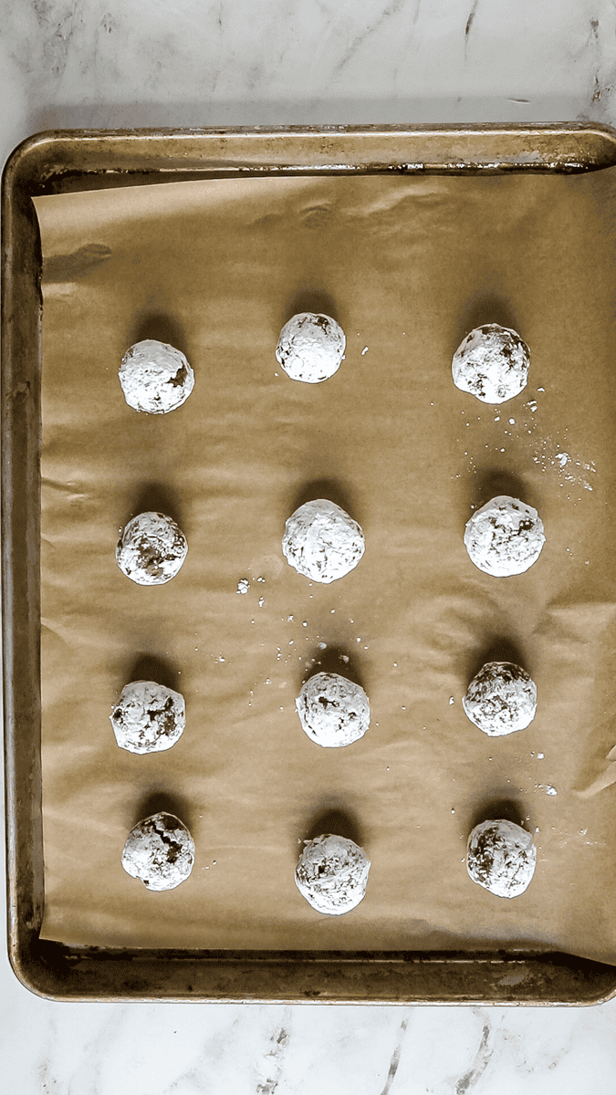12 unbaked chocolate crinkles cookie dough balls rolled in powdered sugar on a baking sheet