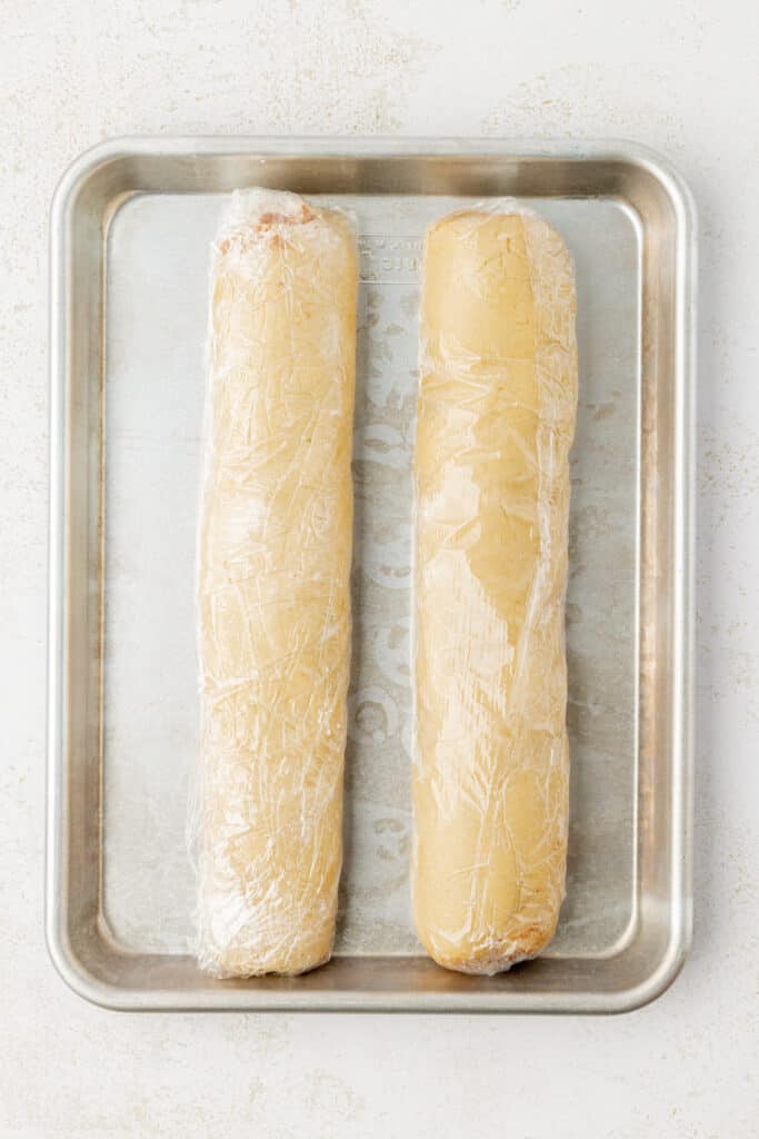 two cookie dough logs wrapped in plastic wrap on a cookie sheet